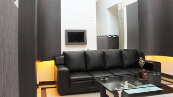 A modern living room with a black leather sofa, a glass coffee table, and a flat-screen TV mounted on the wall.