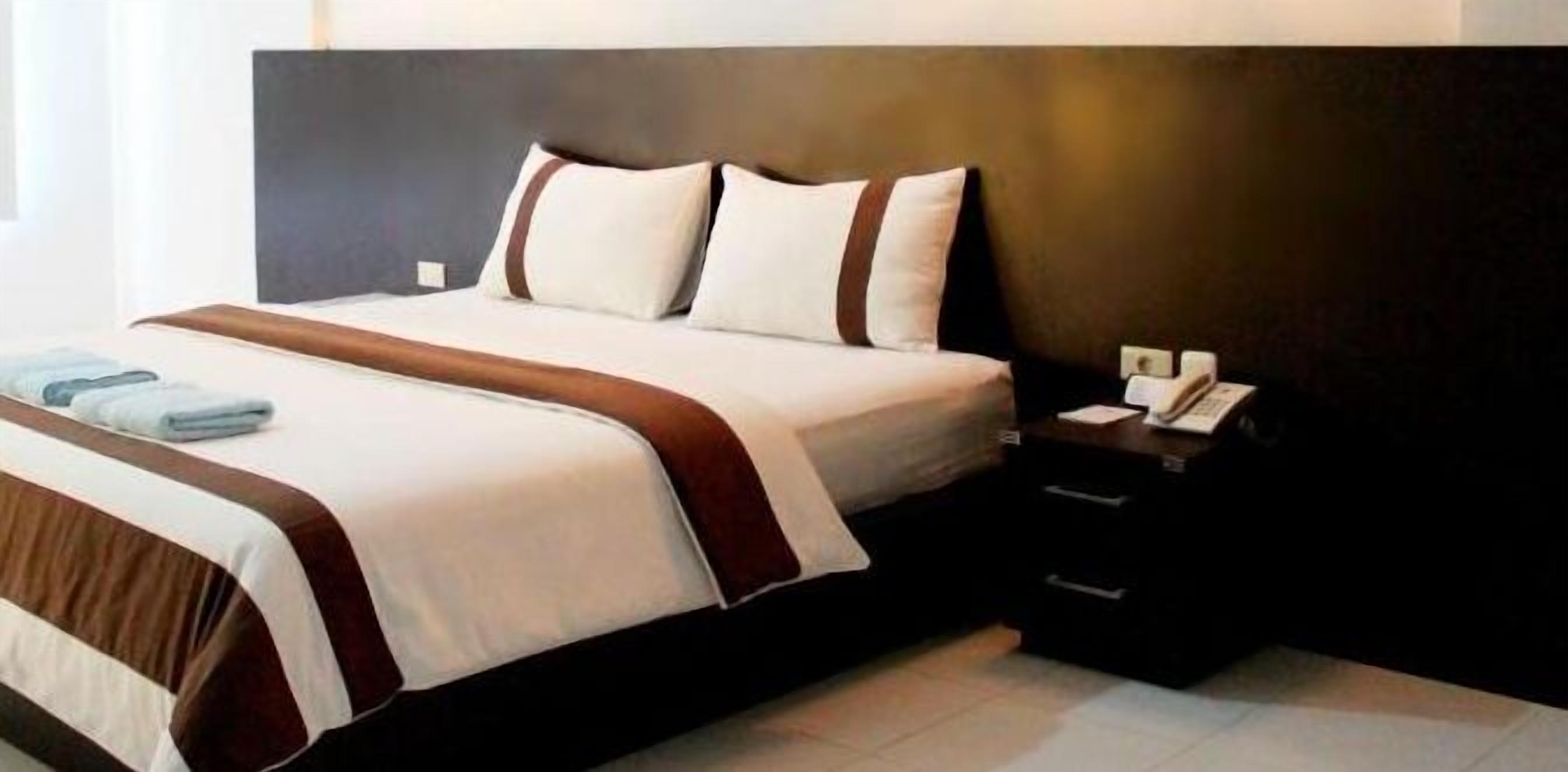 A neatly made bed with white and brown linens, a dark headboard, and a bedside table with a phone and a lamp.