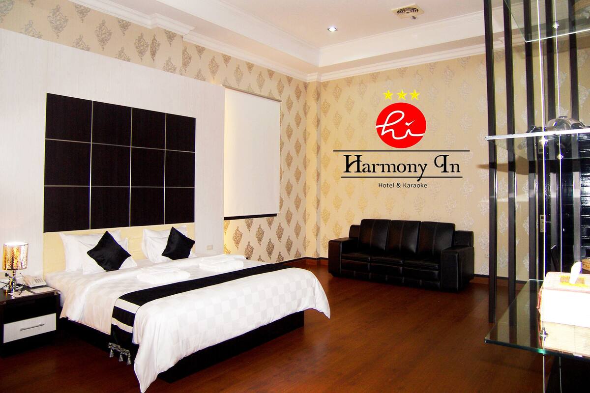 A hotel room with a large bed, a sofa, a glass table, and a wall with the hotel's logo.