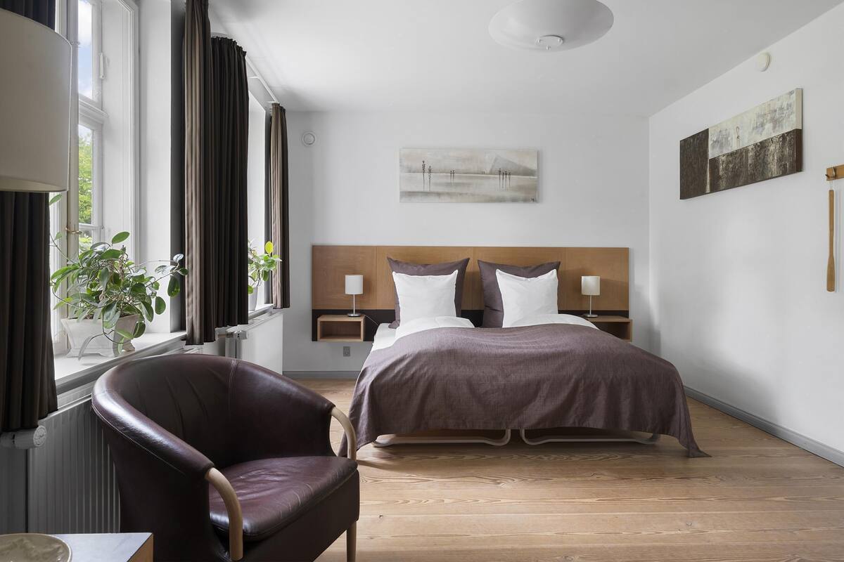 A hotel room with a bed, a chair, a window with curtains, and artwork on the walls.