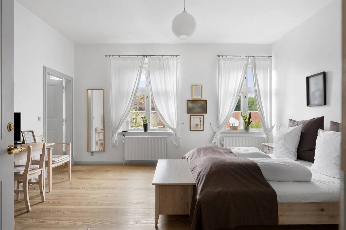 A bedroom with a bed, a desk, a chair, a window with curtains, and a radiator.
