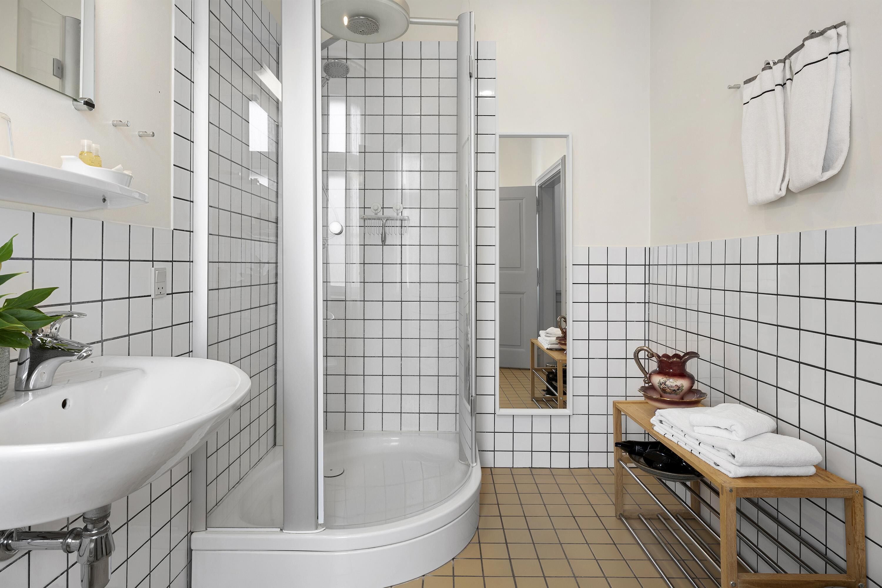 A bathroom with a white sink, a shower, a mirror, and a shelf with towels and a pitcher.