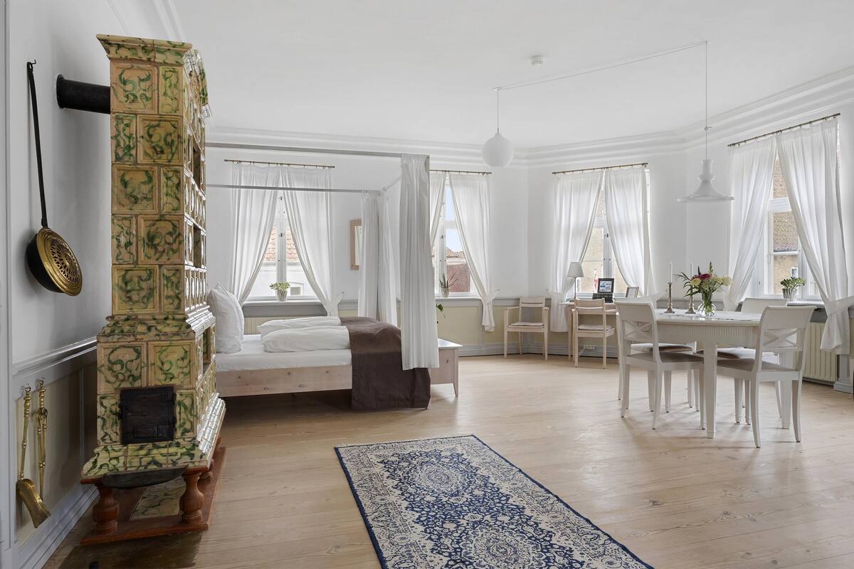 A spacious room with a bed, a dining table with chairs, a large window with curtains, and a decorative column.