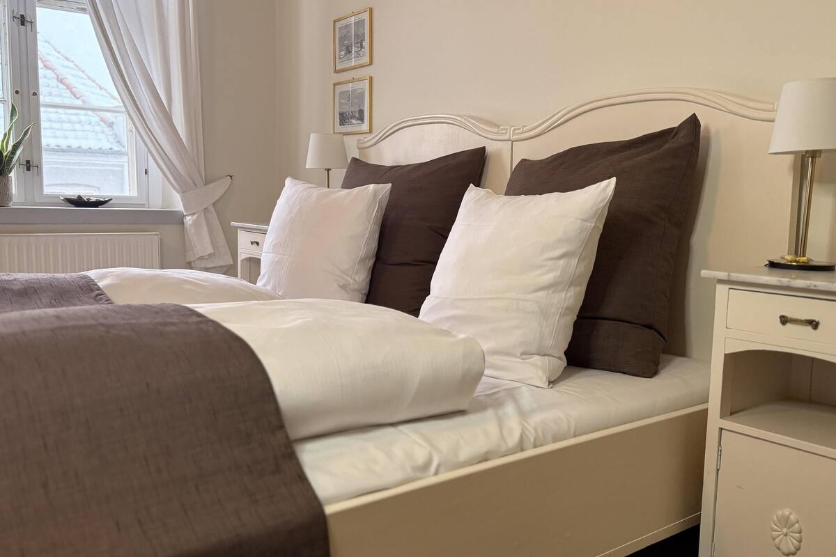 A neatly made bed with white and brown bedding, a bedside table with a lamp, and a window with curtains.