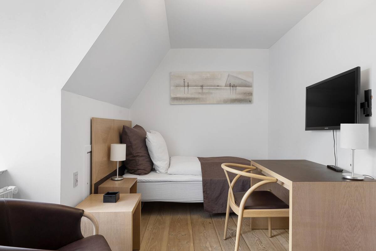 A modern, compact room with a bed, a desk, a chair, a TV, and a painting on the wall.