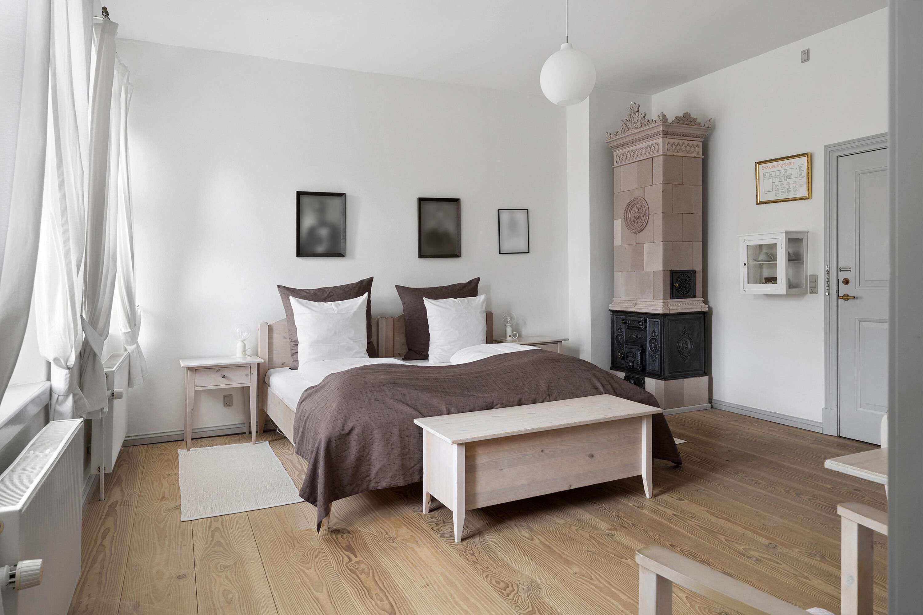 Un dormitorio con una cama, mesitas de noche, un banco, un radiador, una puerta blanca y una chimenea.