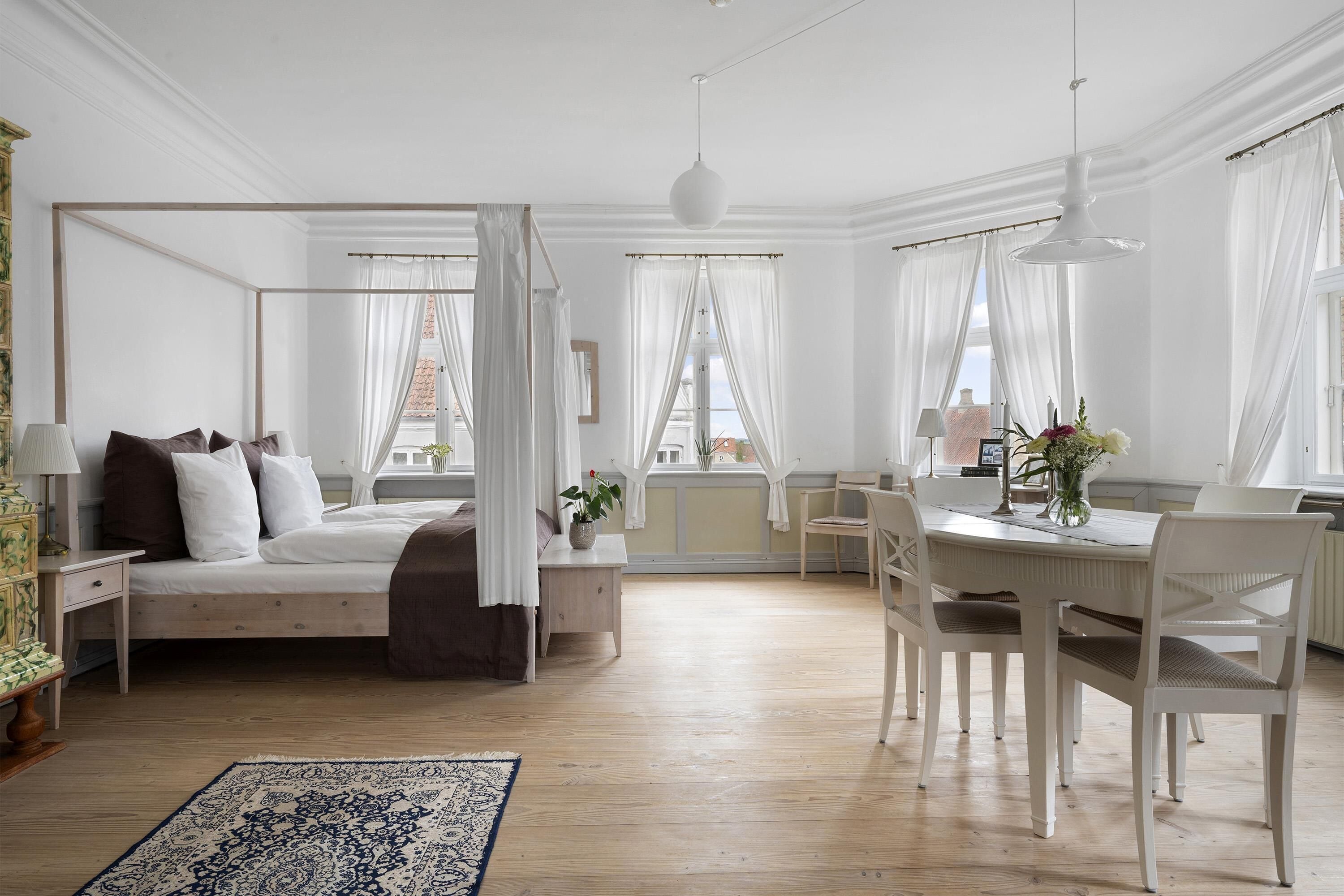 Un dormitorio con cama con dosel, una mesa de comedor con sillas y un amplio ventanal con cortinas.