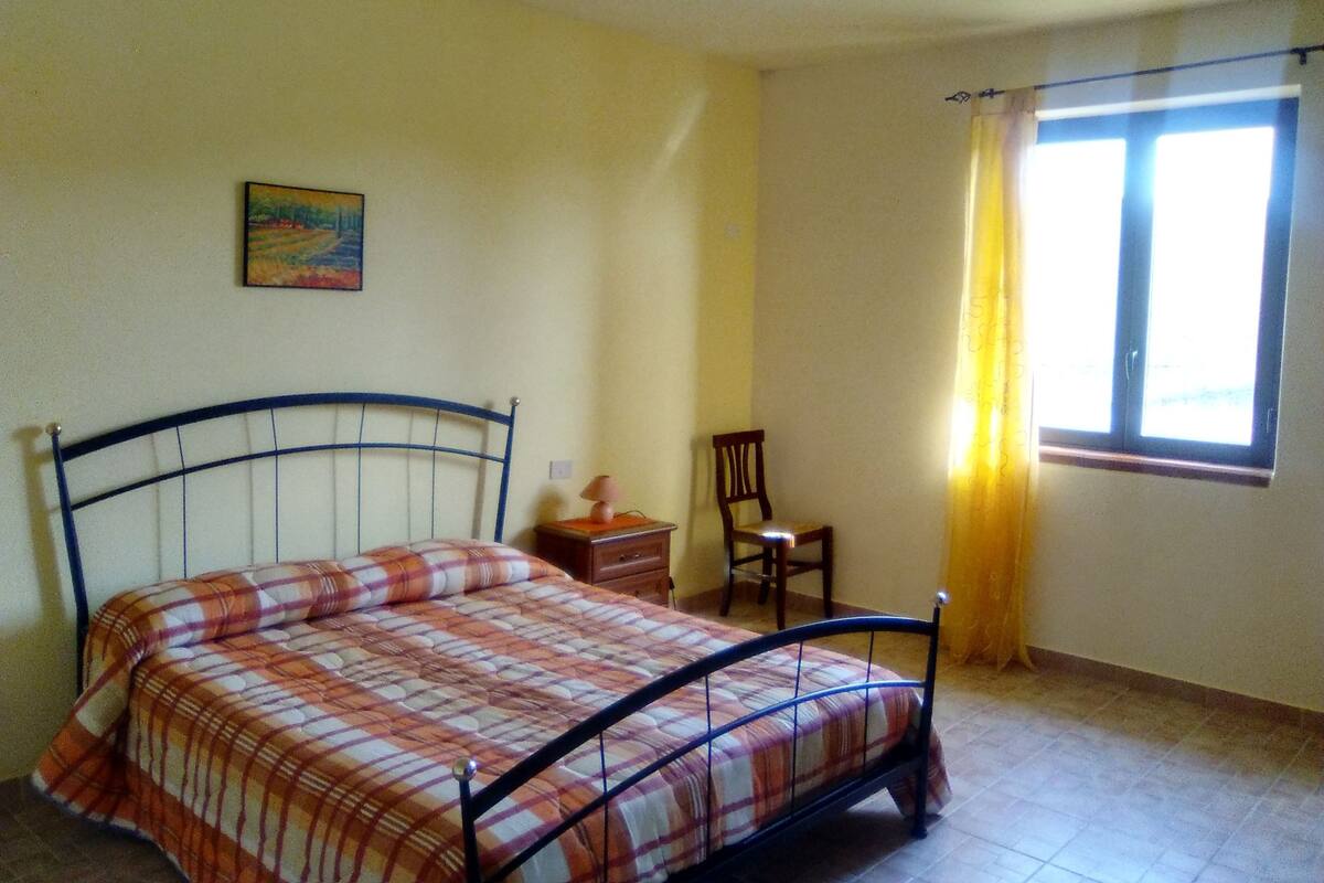 Une chambre à coucher avec un lit, une chaise, une table de chevet, une fenêtre avec des rideaux et un tableau au mur.