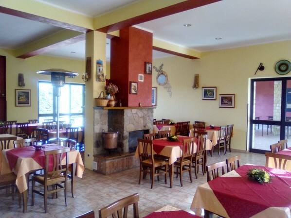 Un espace repas comprenant plusieurs tables et chaises, une cheminée en pierre et une grande fenêtre.