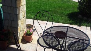 Un patio con un pilar de piedra, una mesa y sillas de metal, una piscina y un edificio con techo de tejas al fondo.