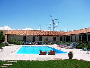 Un espace piscine avec des chaises longues et un bâtiment doté d’un toit recouvert de tuiles.