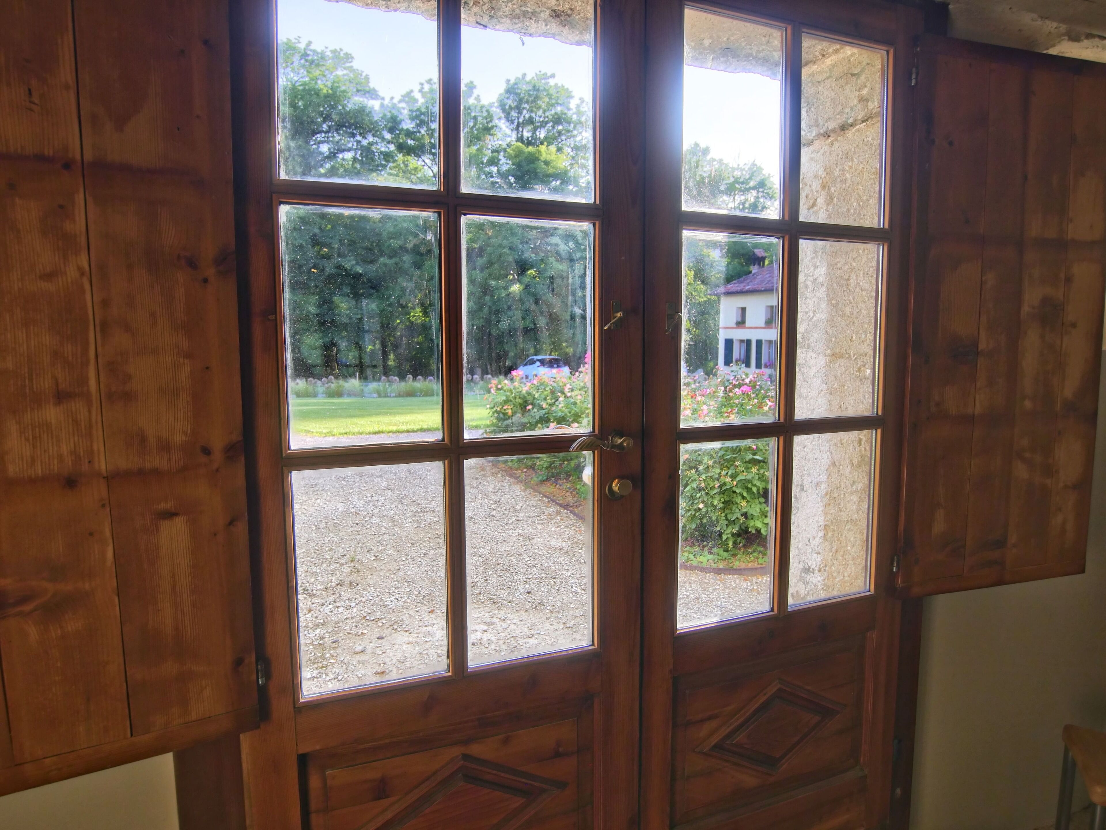 Una porta in legno con sei vetri che si apre su un giardino con un vialetto sterrato e un edificio sullo sfondo.