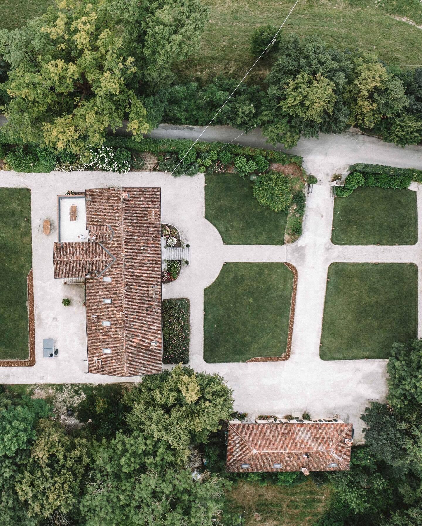 An aerial view of a property with a large brick building, a patio area with outdoor furniture, and a well-maintained lawn with geometric garden beds.