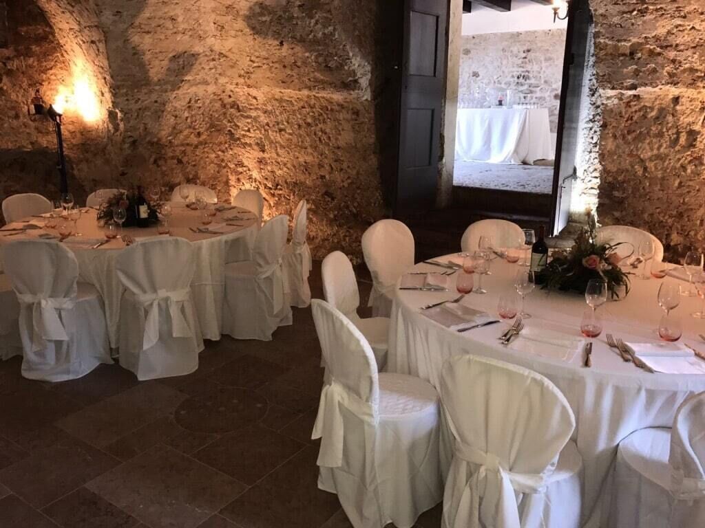 A rustic dining area with stone walls and white tablecloths. Tables are set with wine glasses, cutlery, and flowers.