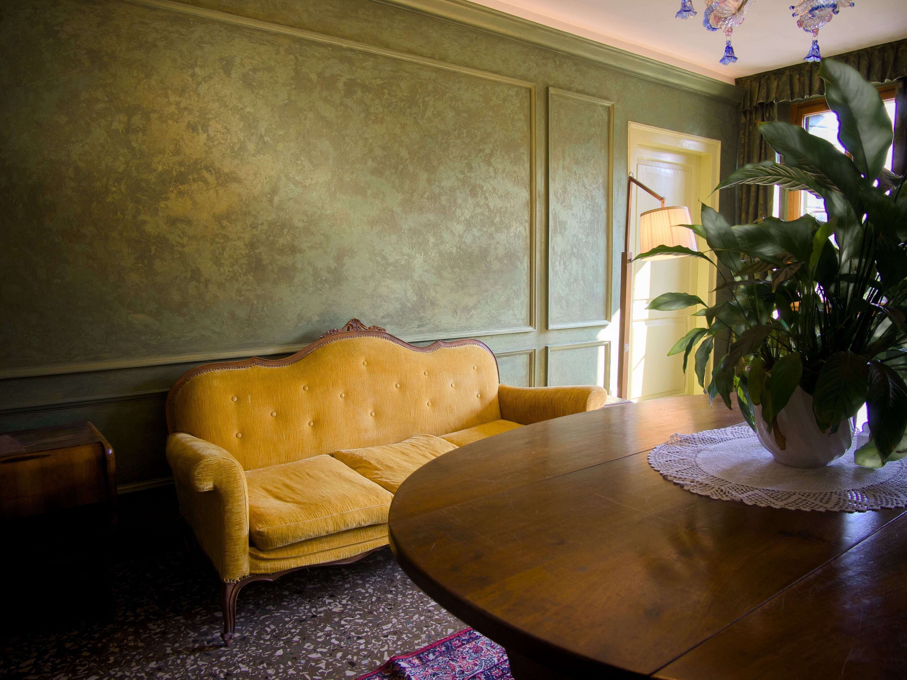 Una habitación con una mesa redonda de madera, un sofá amarillo, una planta en maceta y un tapete estampado.