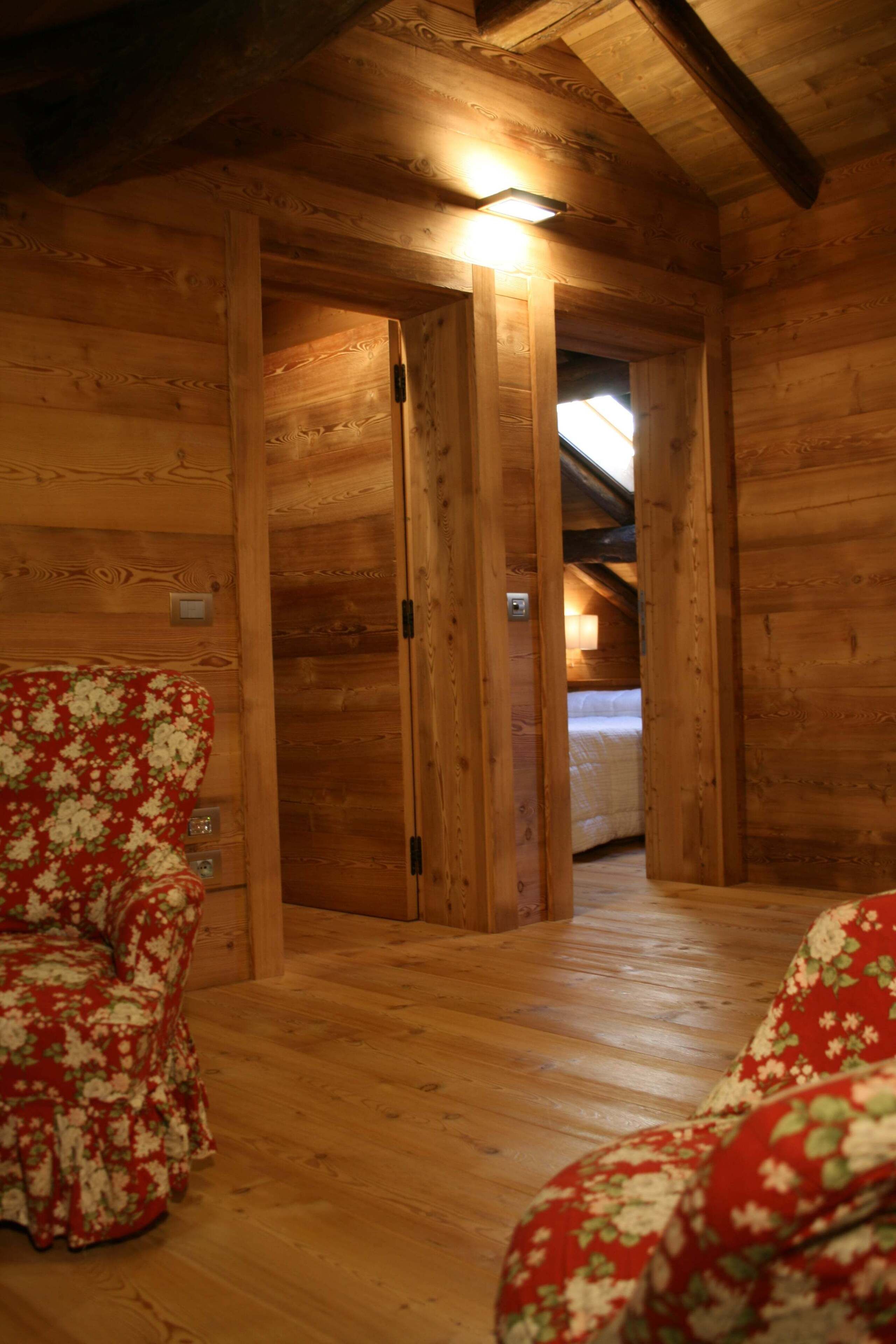 Interior de una cabaña de madera con una cama acogedora, techo de madera y una silla roja con estampado floral.