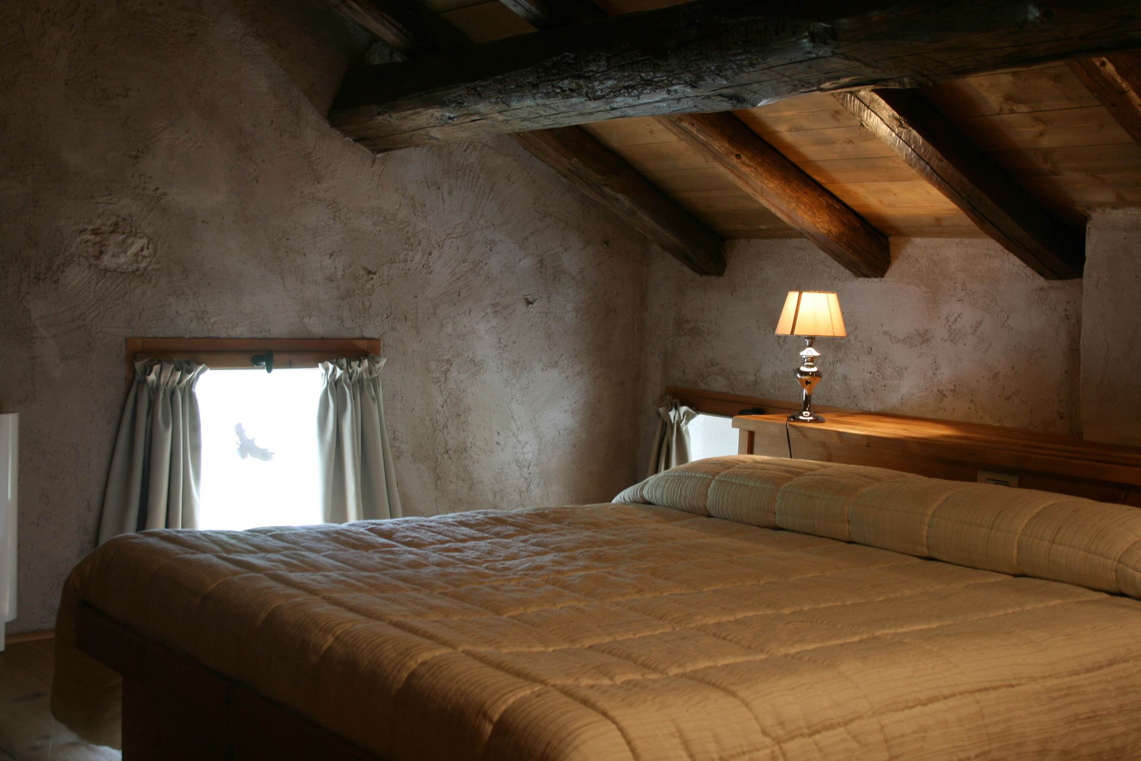 A bedroom with a large bed, a nightstand with a lamp, a window with curtains, and exposed wooden beams on the ceiling.
