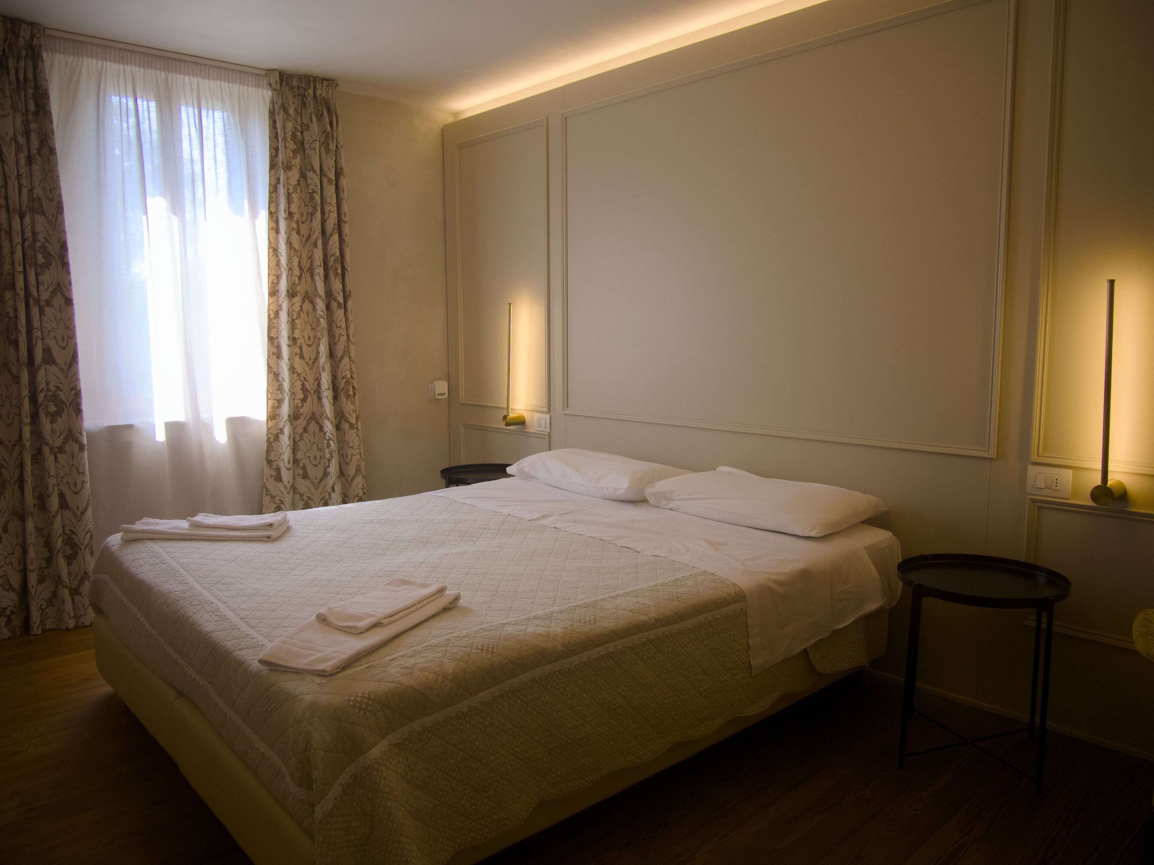Un lit bien fait, avec du linge de lit blanc et une serviette pliée, une table de chevet avec une lampe et une fenêtre ornée de rideaux légers.