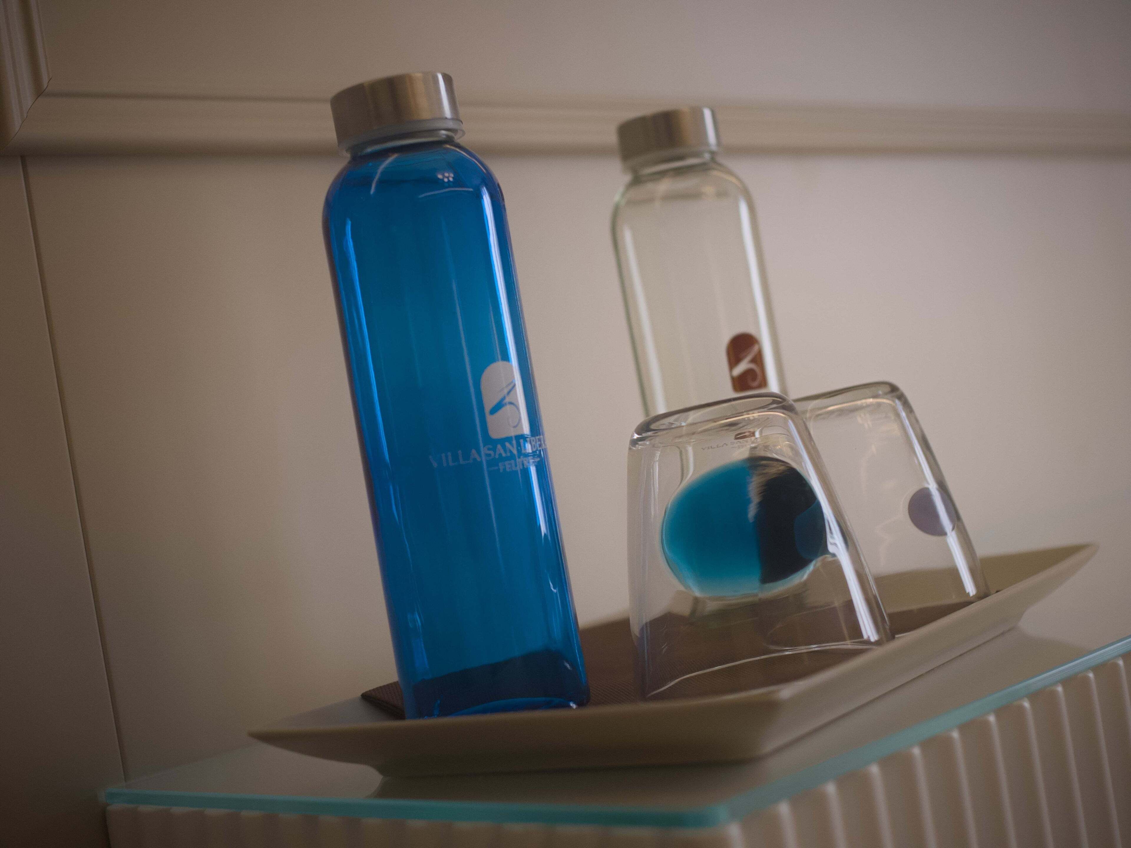 Une étagère en verre sur laquelle reposent deux bouteilles, l’une bleue et l’autre transparente, sur une surface blanche.