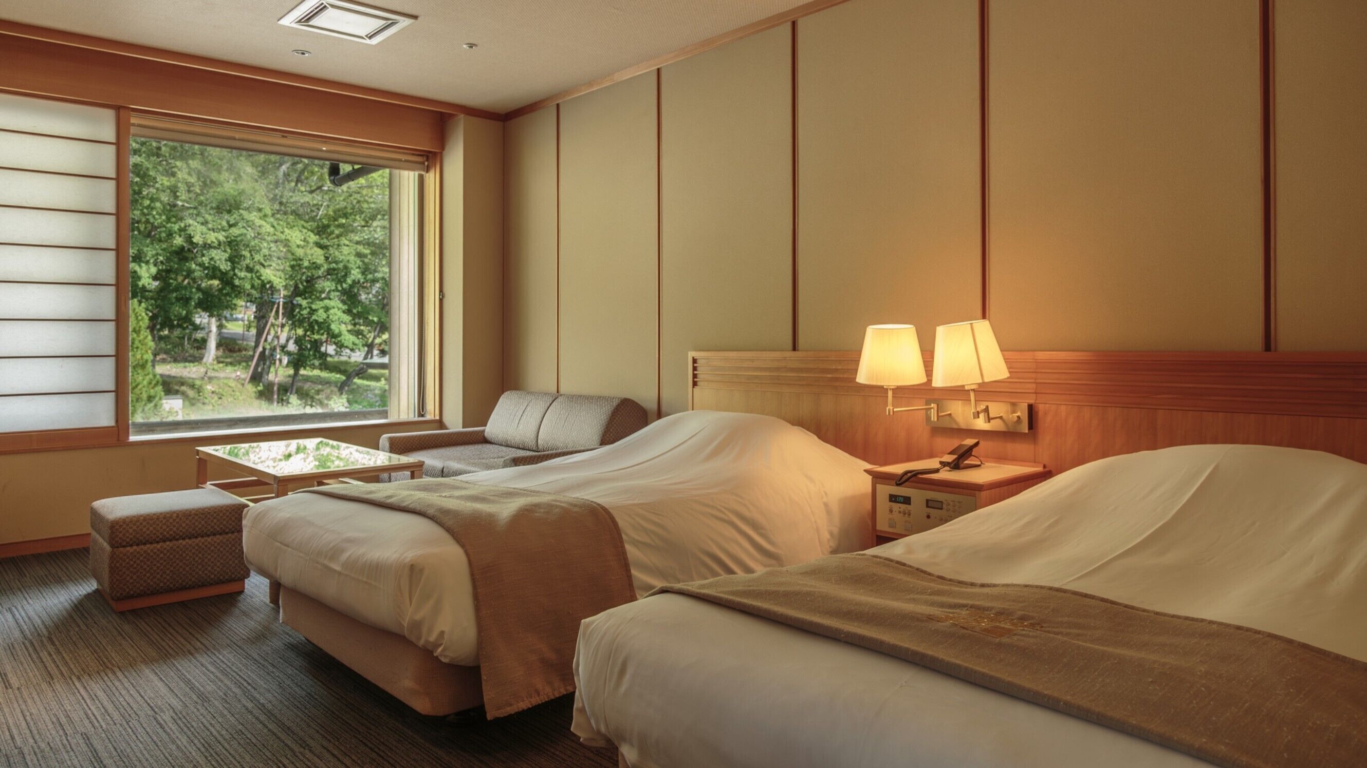 A hotel room with two beds, a sofa, a small table, and a window with a view of trees.