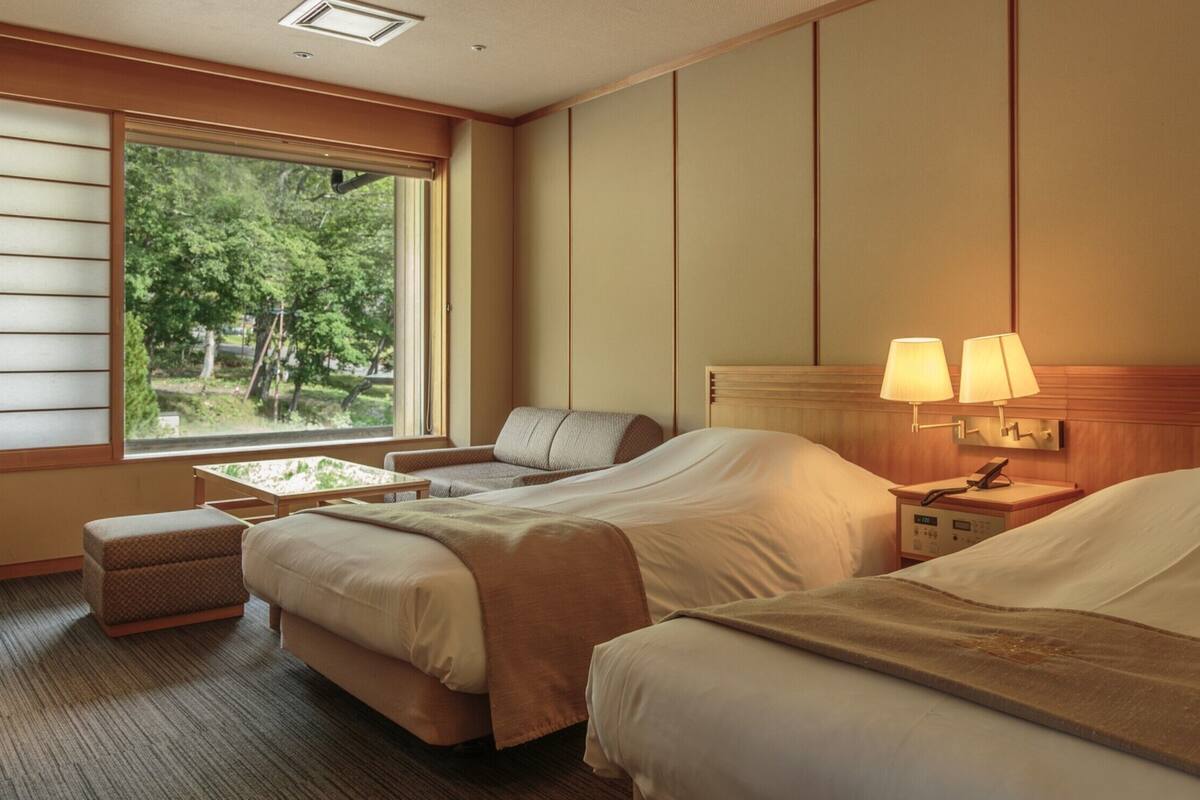 A hotel room with two beds, a sofa, a small table, and a window with a view of trees.