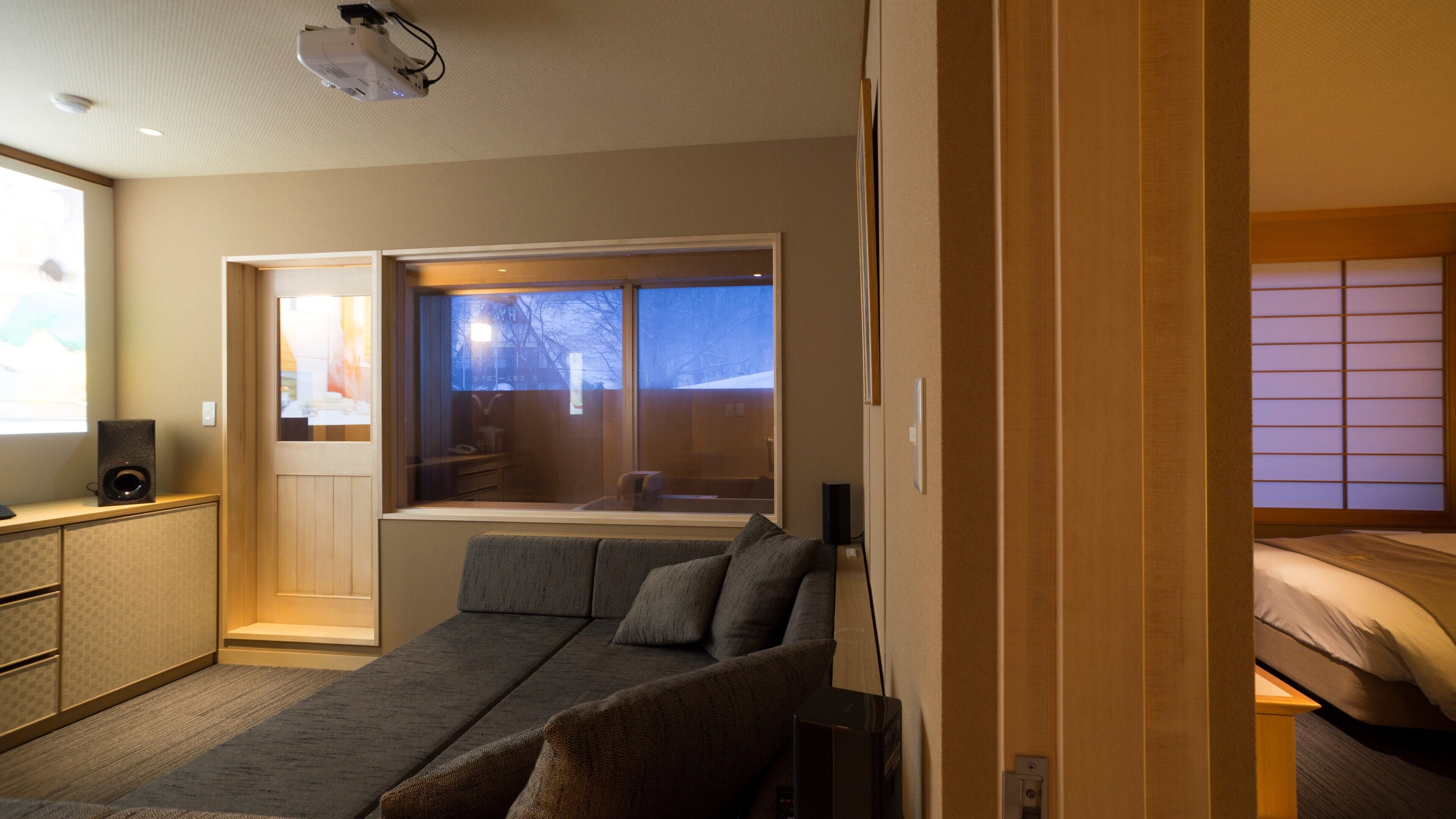A modern hotel room with a sofa, a bed, a window with a view, a projector, and a speaker.