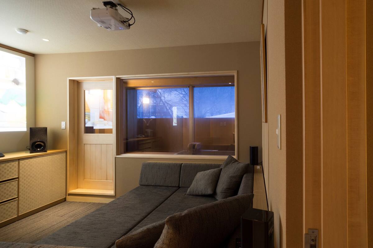 A modern hotel room with a sofa, a bed, a window with a view, a projector, and a speaker.