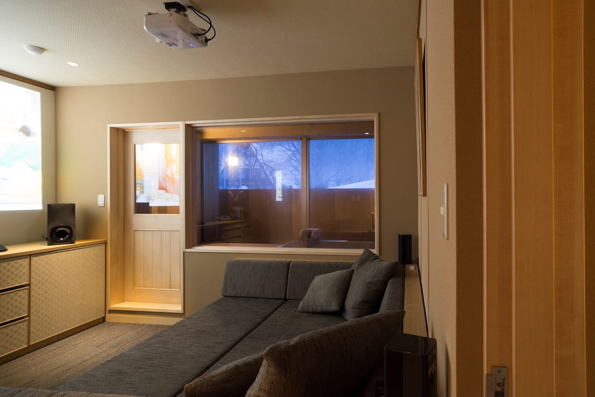 A modern hotel room with a sofa, a bed, a window with a view, a projector, and a speaker.