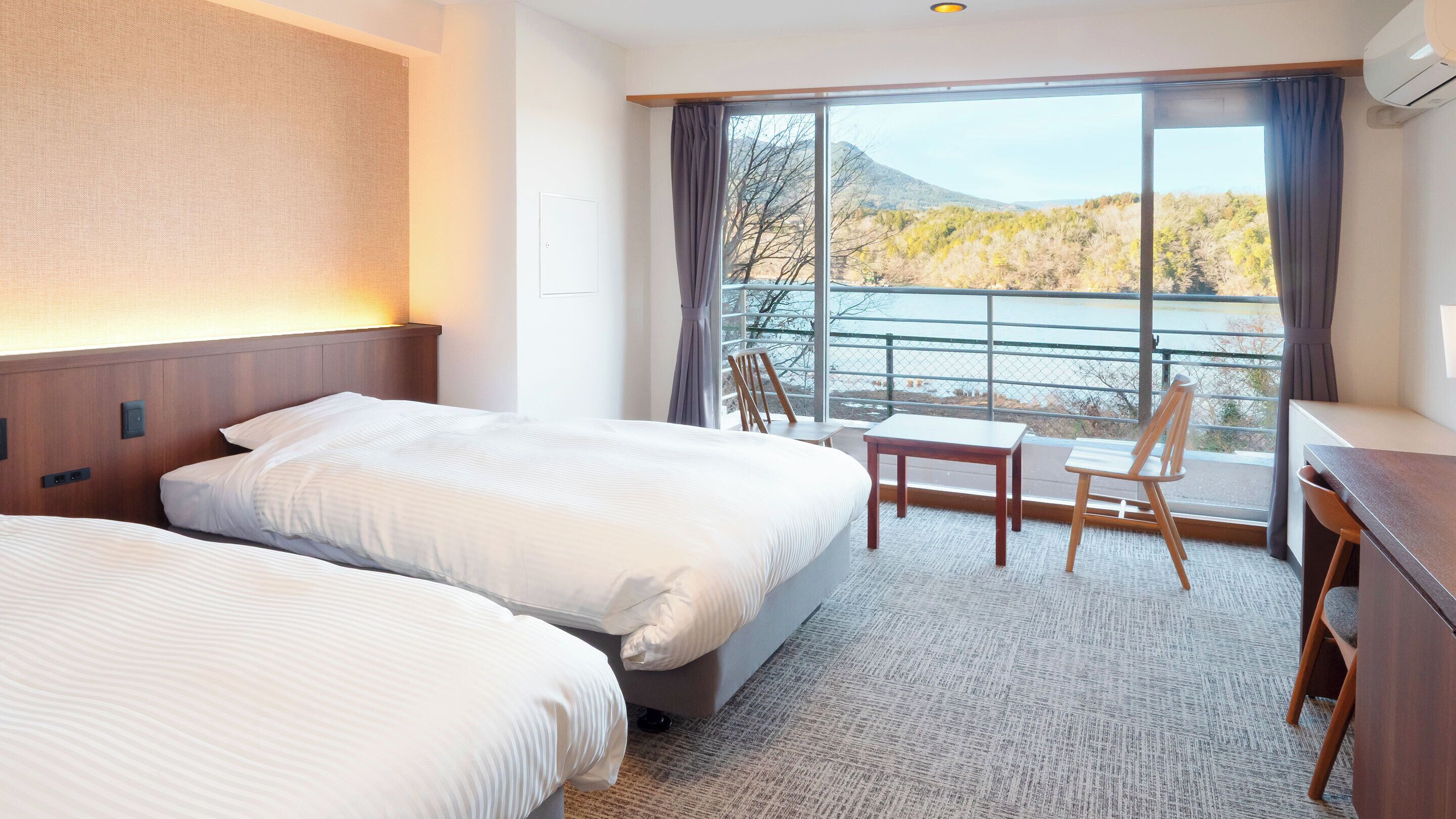 A hotel room with two beds, a desk, and a chair, with a view of a lake and mountains.