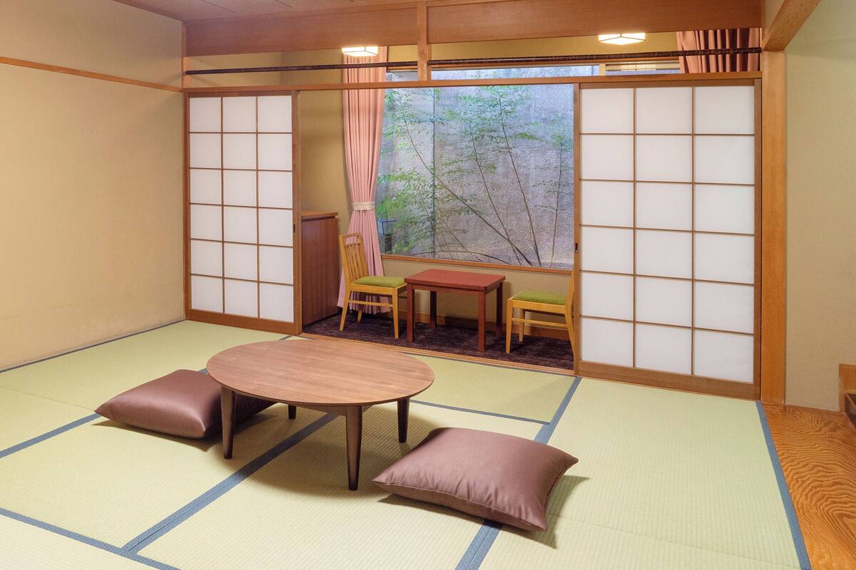 A tatami mat room with a low table and two cushions, a sliding door with a view of trees, and a small table with a chair.