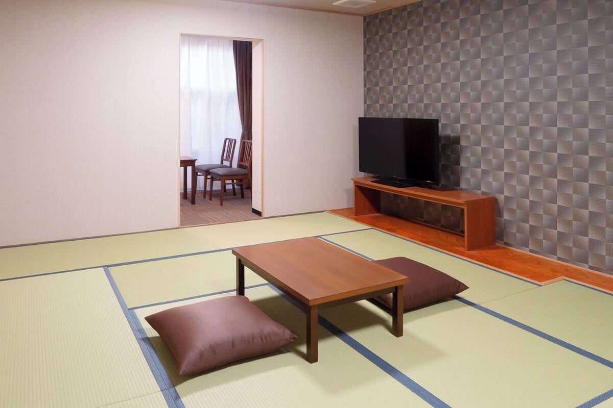 A traditional Japanese-style living room with tatami flooring, a low wooden table, cushions, and a television on a wooden stand.