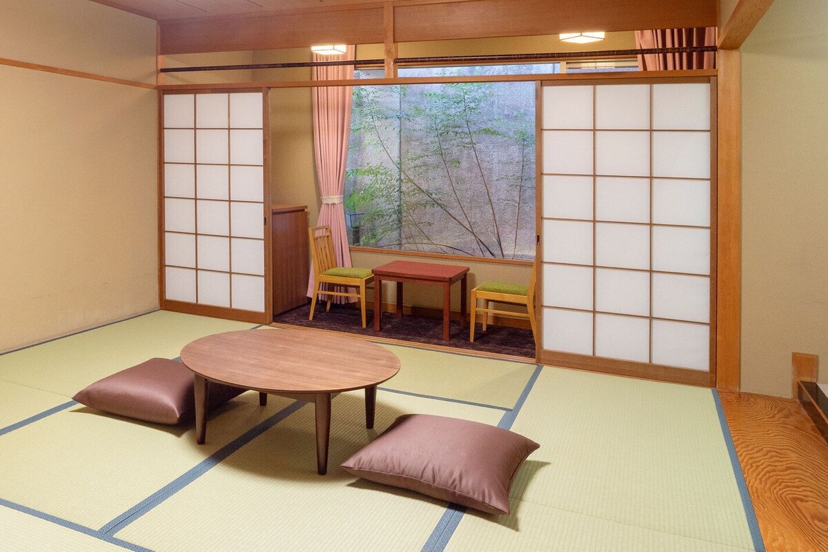 Ein Raum mit Tatami-Matten, einem niedrigen Tisch, Kissen, einer Schiebetür mit Blick und einem kleinen Tisch mit Stühlen.