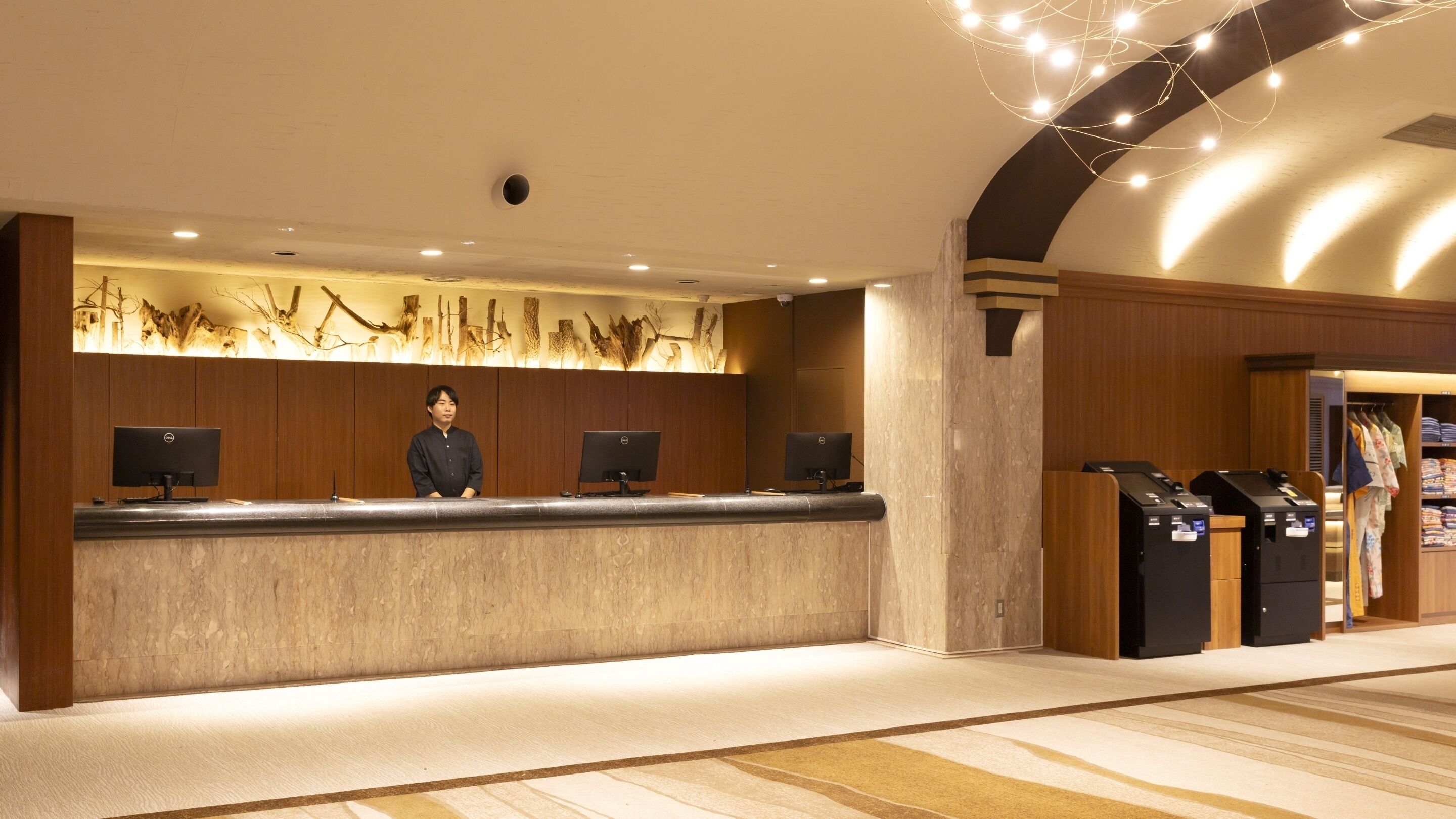 A hotel reception desk with a receptionist, three computer monitors, a decorative wall, and a luggage storage area.
