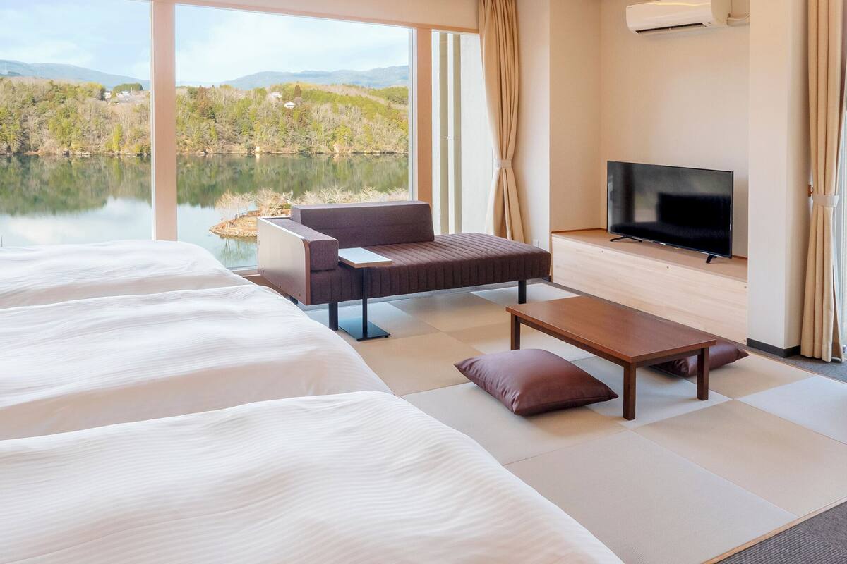 A modern hotel room with a large bed, a sofa, a TV, and a view of a lake and mountains.