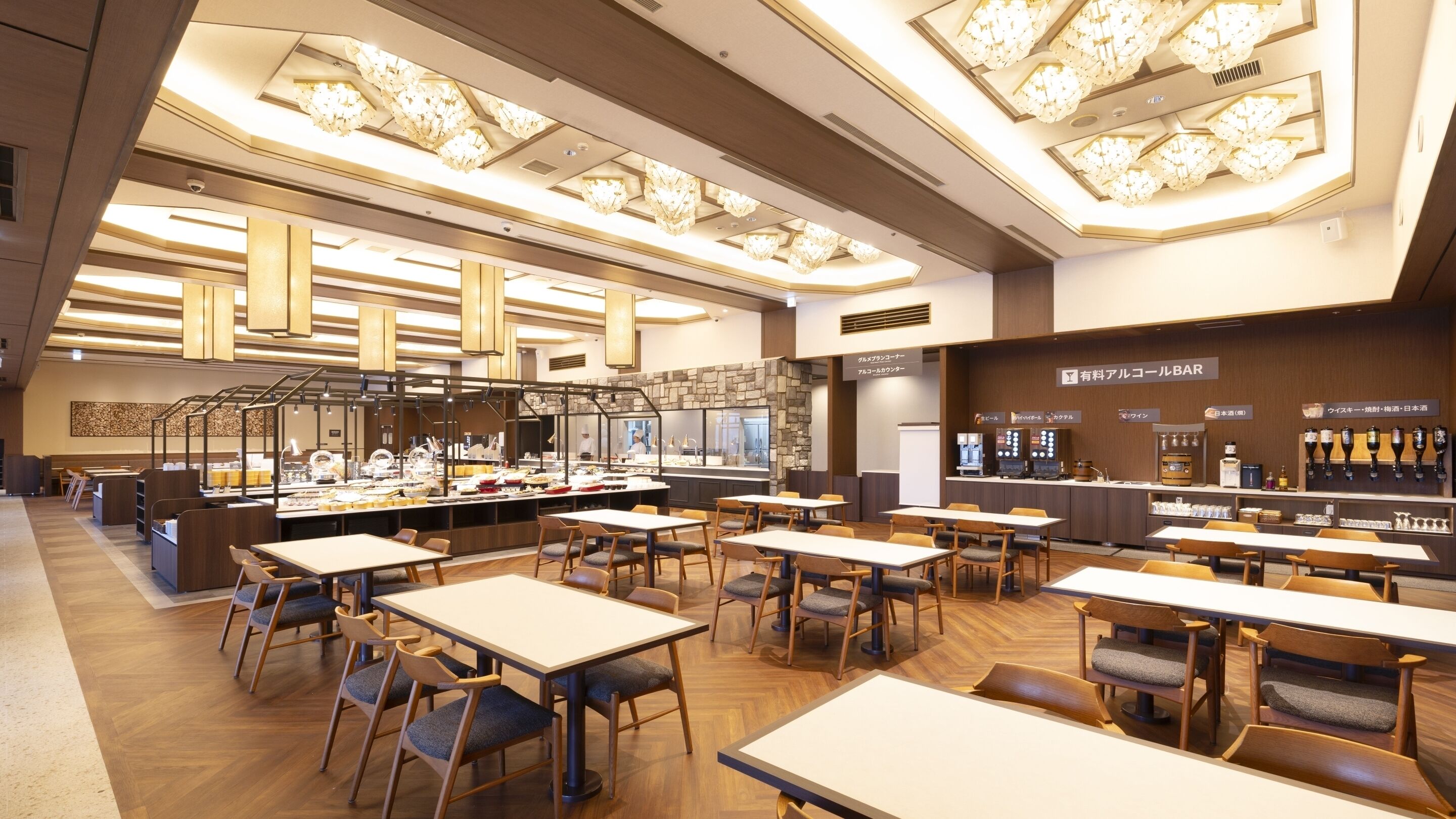Ein modernes Restaurant mit mehreren Tischen und Stühlen, einer Bar mit verschiedenen Getränken und einer Küche im Hintergrund.