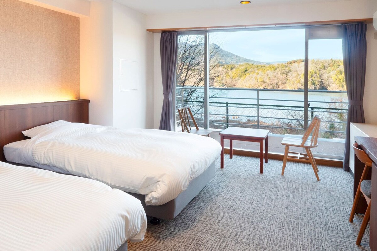 Ein Hotelzimmer mit zwei Betten, einem Schreibtisch, einem Stuhl und einem großen Fenster mit Blick auf einen See und Berge.