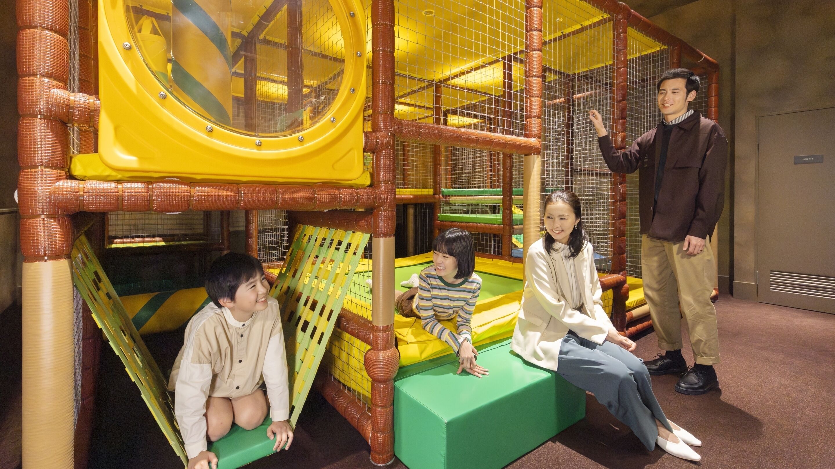 Un espace de jeux avec un toboggan jaune, des structures de jeux vertes et jaunes, et un enclos protégé par un filet.