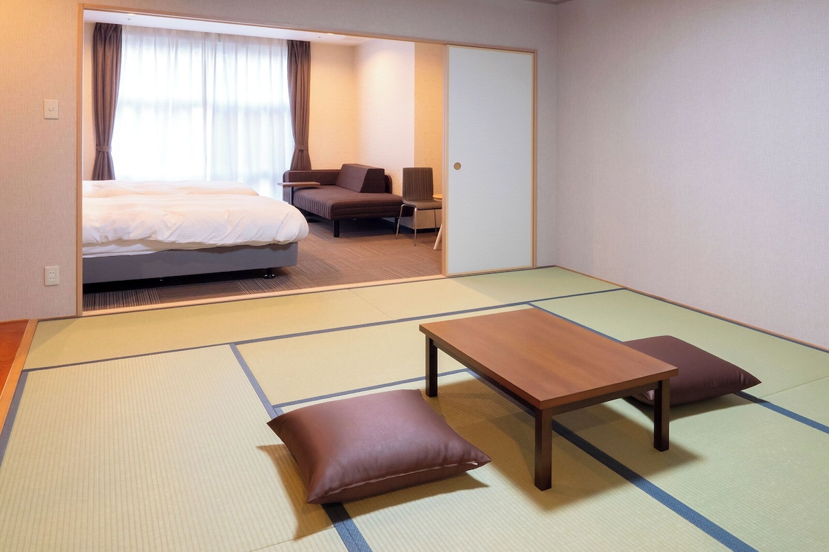 Ein Hotelzimmer mit Tatami-Boden, einem Bett, einem Sofa, einem Tisch und einem Stuhl.