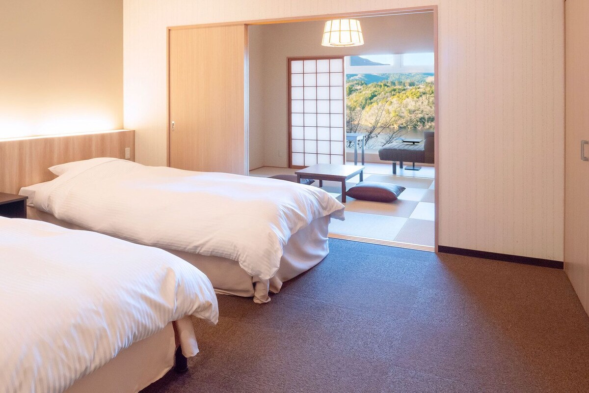 Ein Hotelzimmer mit zwei Betten, einem kleinen Tisch und Blick auf einen Garten durch eine Schiebetür.