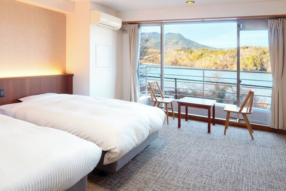 A hotel room with two beds, a table, chairs, a view of a lake, and a mountain in the background.