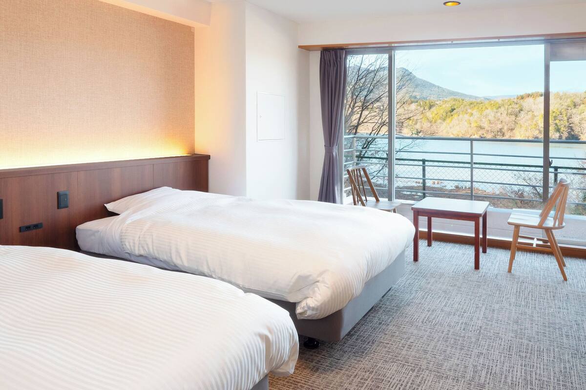 A hotel room with two beds, a desk, and a chair, with a view of a lake and mountains.