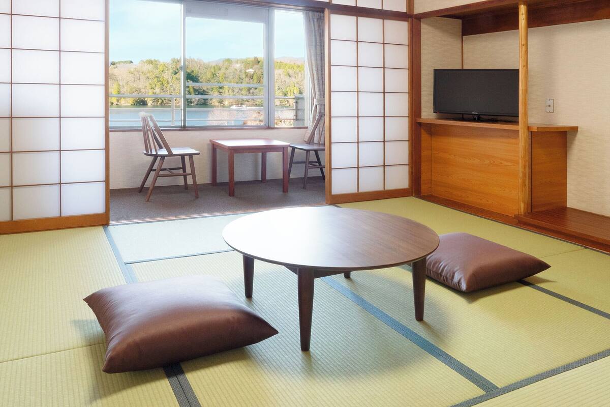 A traditional Japanese room with tatami flooring, a low table, cushions, a TV on a wooden shelf, and a view of a lake through a sliding door.