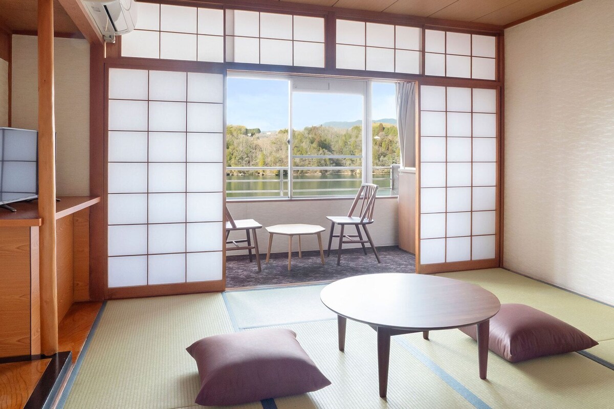 Ein traditioneller japanischer Raum mit Tatami-Boden, einem niedrigen Tisch, zwei Kissen und einem Fernseher auf einer Holzablage.