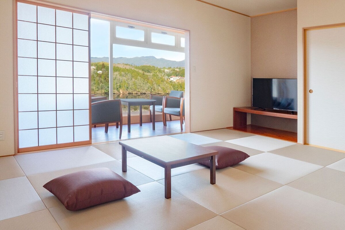 Ein Zimmer mit Tatami-Boden, einem niedrigen Tisch mit Kissen, einem Fernseher auf einem Holzständer und einer Schiebetür zu einem Außenbereich mit Stühlen.