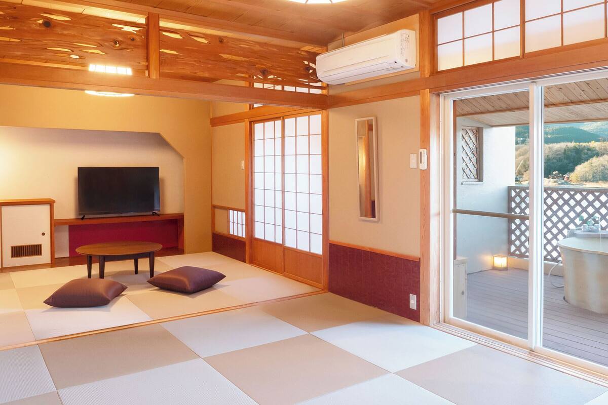 A traditional Japanese-style room with tatami flooring, a television, a table, and a sliding glass door leading to an outdoor area.