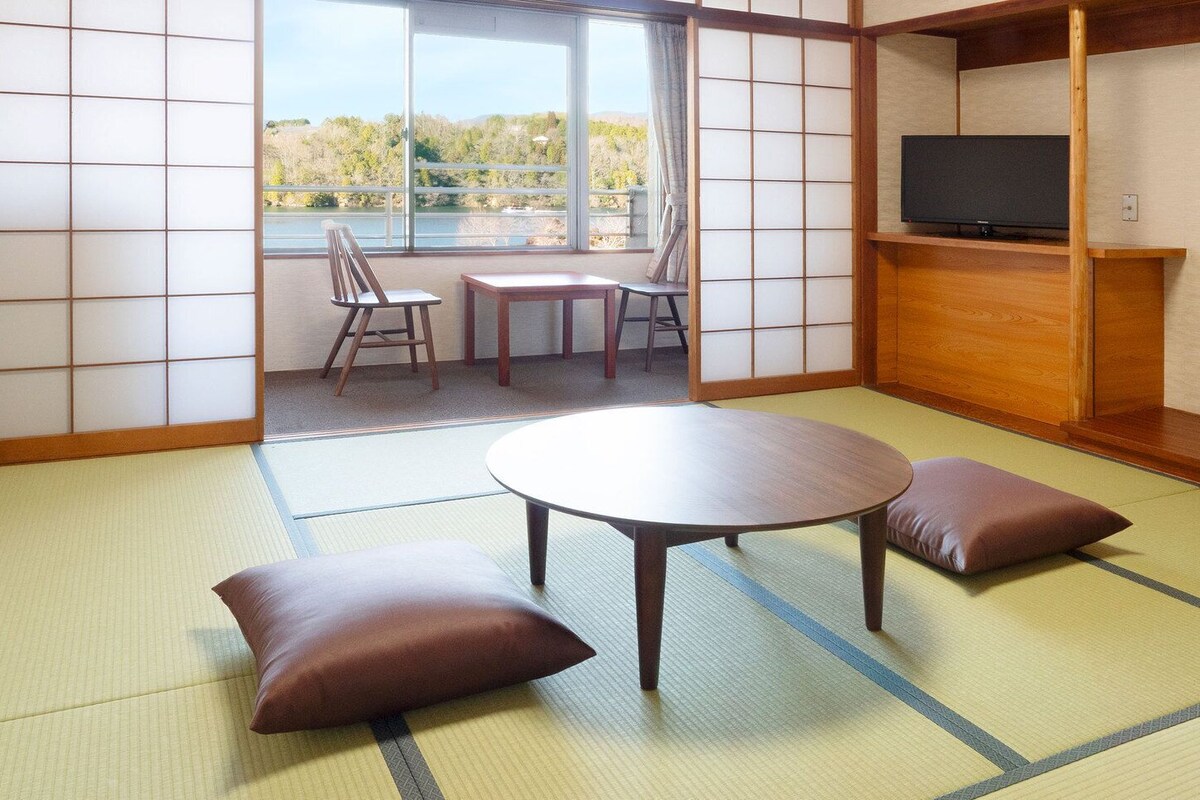 Ein Raum mit Tatami-Matten, einem runden Tisch, zwei Kissen, einem Fernseher auf einem Holzregal und Blick auf einen Balkon mit Stühlen.