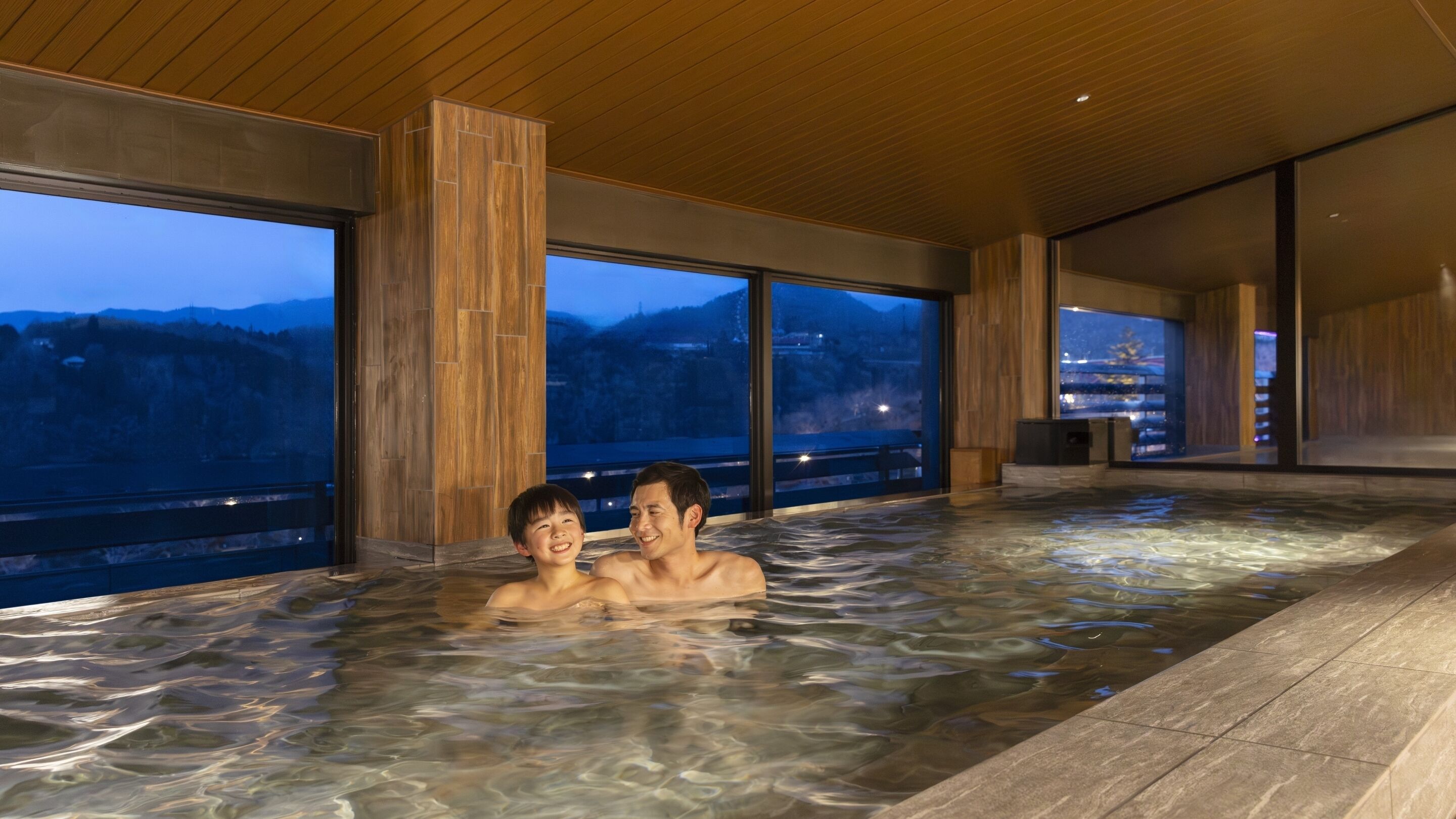 A hot tub with two people, large windows, and a view of mountains.
