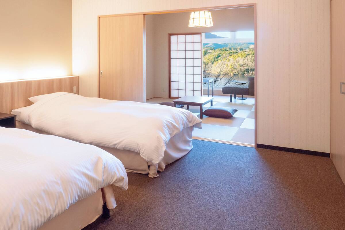 A hotel room with two beds, a small table, and a view of a garden through a sliding door.