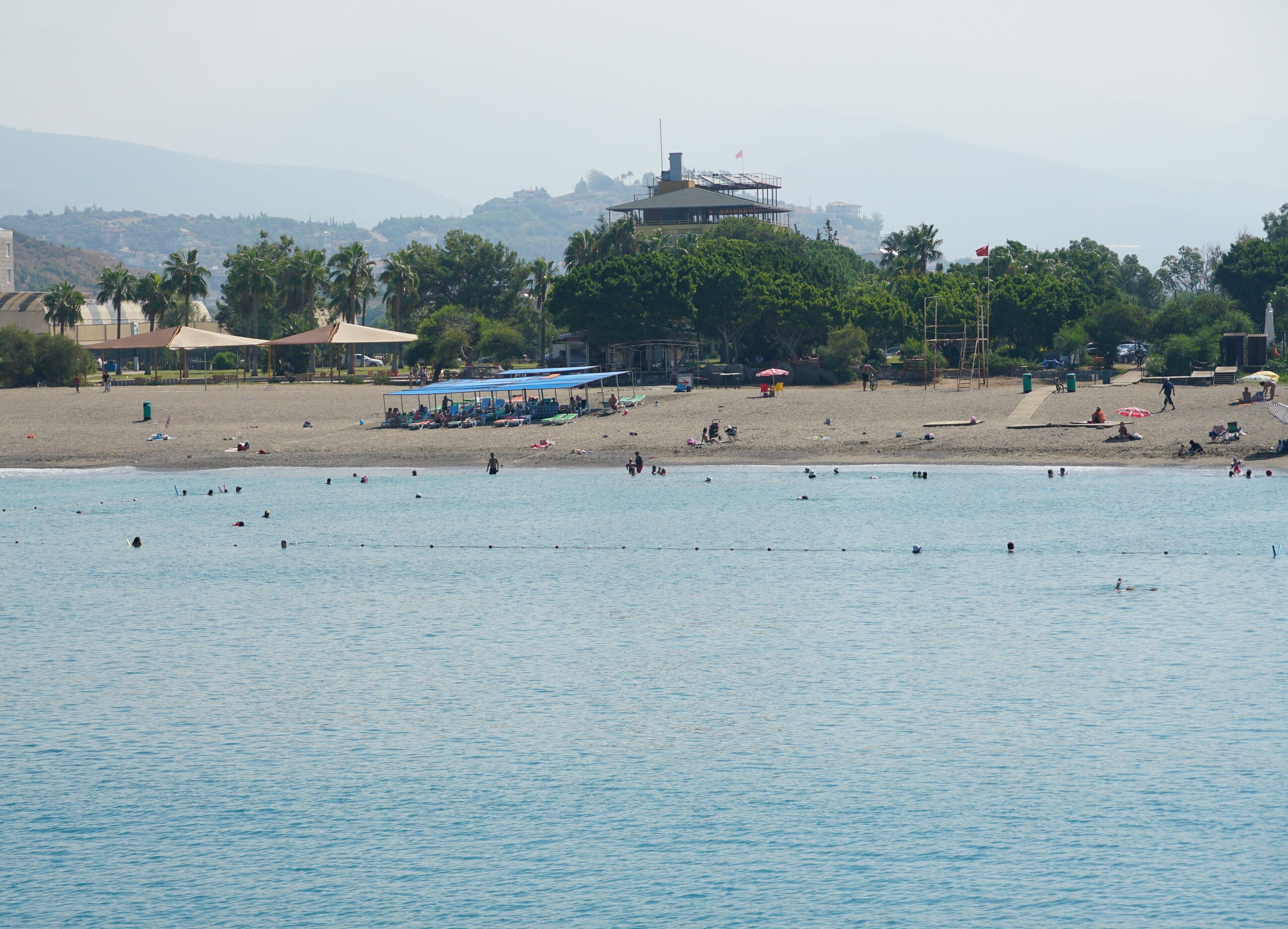 Une plage où l’on voit des gens nager et bronzer, une zone abritée avec des parasols, et un bâtiment en arrière-plan.