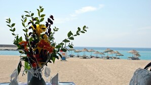 Une table dressée pour deux personnes, agrémentée d’un vase de fleurs, avec une vue sur la plage et des parasols en arrière-plan.